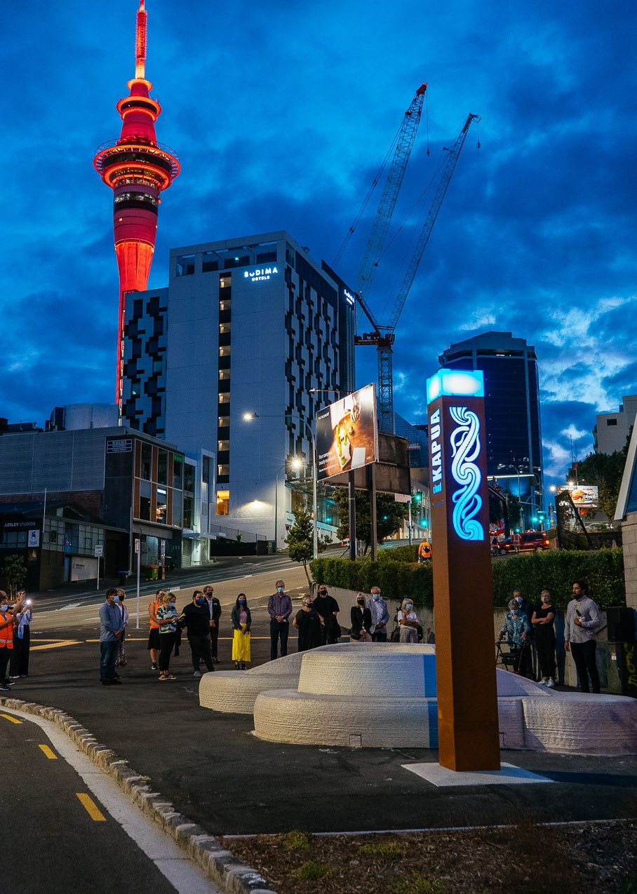 We see the corner of Wellesley and Sale street at dawn; people are gathered around a public artwork with a light installation next to it. On this is the word "kapua". This is the blessing of the artwork where people can sit and reflect. IN the background is the Auckland skyline with hotels, cranes and the sky tower.