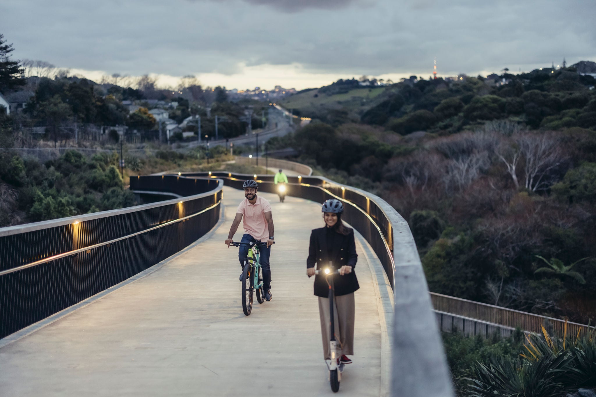 People riding their scooters and bikes home from a long day at work on the Te Ara Ki Uta Ki Tai St Johns Road to Ōrākei Basin Shared Path. It is a beautifully crafted pathway with wooden rails and downlighting for safety, and the natural environment of the Ōrākei Basin is all around.