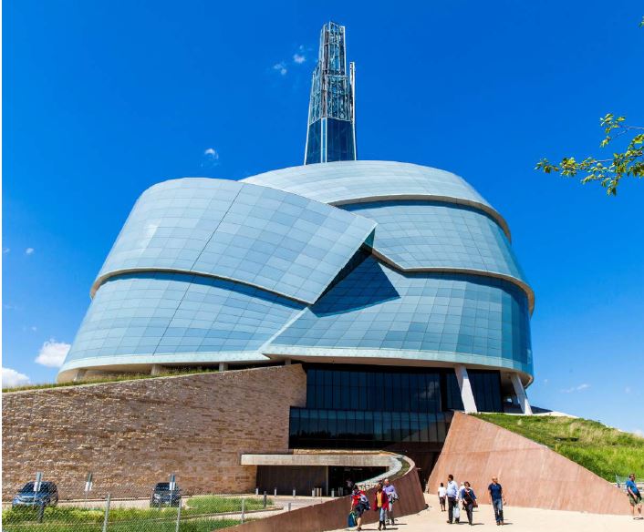 It's a blue sky day and the Canadian Museum for Human Rights sits before us. A light pathway arcs from the right of the picture to the entrance on the bottom level. This level is made of stone and the top levels of the structure bulge to the sky, encased in grey-blue glass. This glass structure is made of bended plates, almost like an armadillo, with a glass spire rising above. People walk in and out of the entrance below, they look tiny in comparison to the building. 