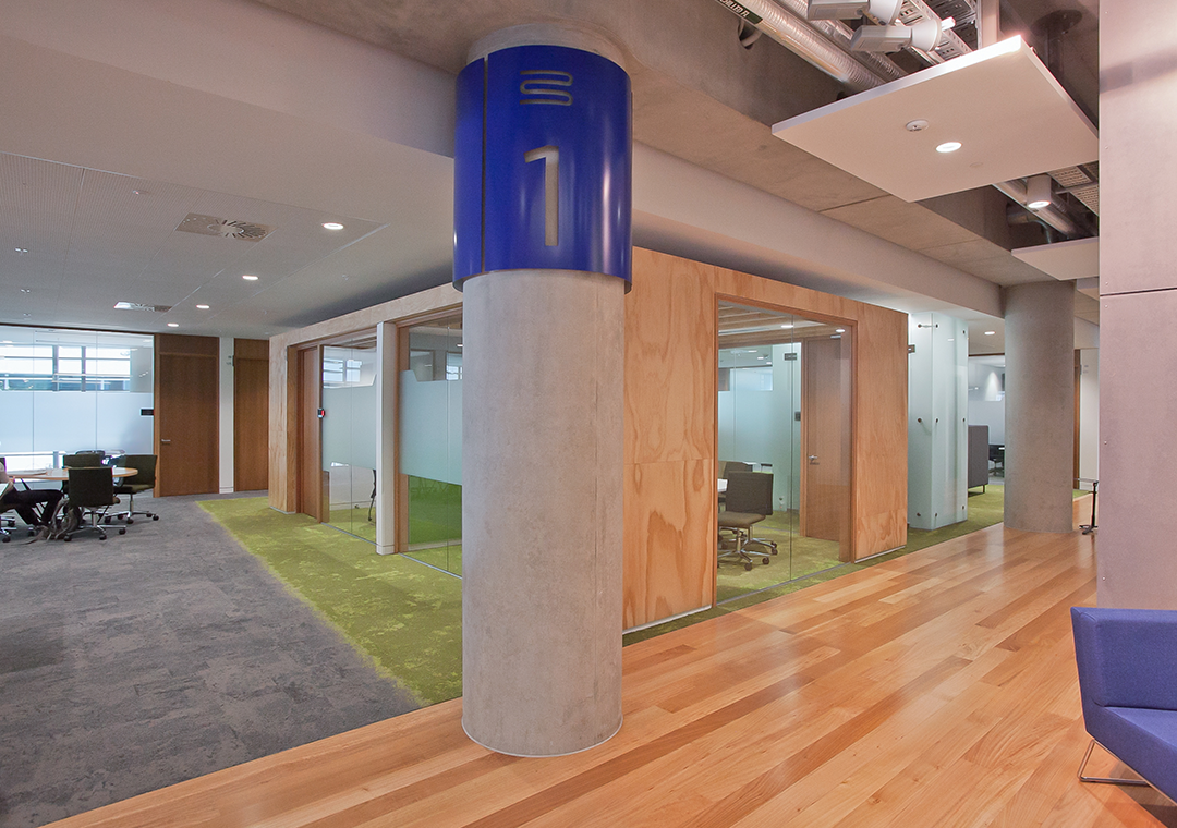 Different types of spaces can be marked by material and colour changes. In this case, a wooden floor is used for circulation areas, grey carpet is for open-plan office space, and green carpet marks private meeting rooms. Colour coded floor numbering can assist with wayfinding.