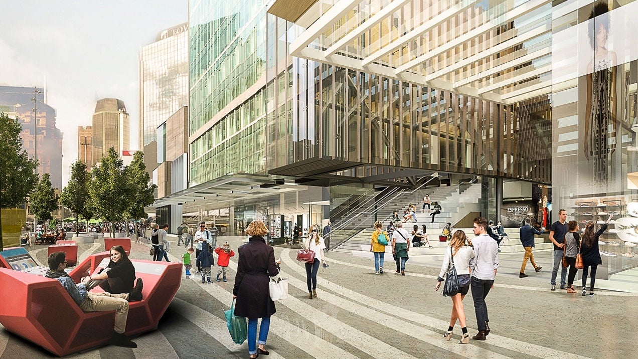 We see a first person view of Commercial Bay plaza, looking south. It is daytime, and people are out sitting on benches enjoying each other's company, while others walk with shopping bags. The glass facade of Commercial Bay towers over the people, hemmed in by trees. We see Queen street in the background.