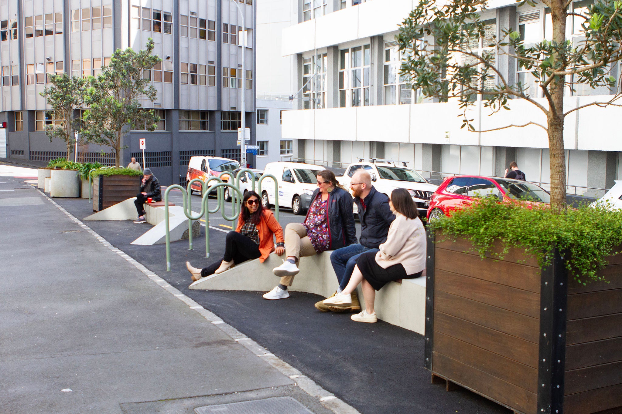 A diverse group of five people sit on the concrete seating amongst the planter boxes and bike racks at St Paul's street in downtown Auckland. The trees are hemmed by native climbing plants.