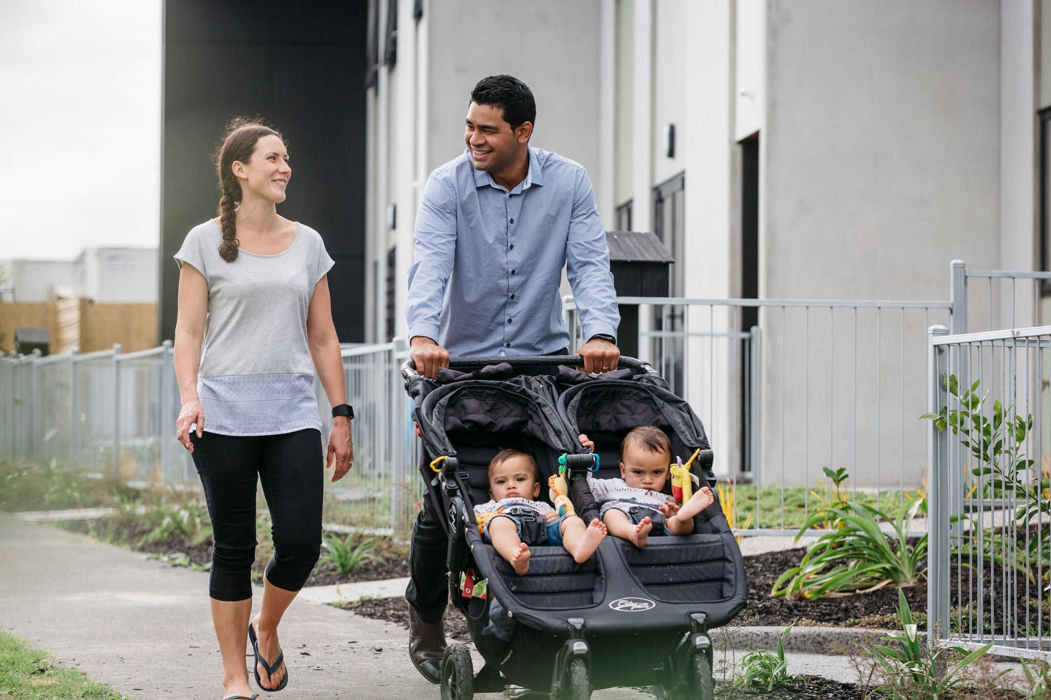 A mum and dad share a smile while walking down a footpath in the evening after work. Dad pushes a stroller with their twin babies. They are walking down a footpath in front of terraced homes. There are front gardens and low fences between them and the gardens. 