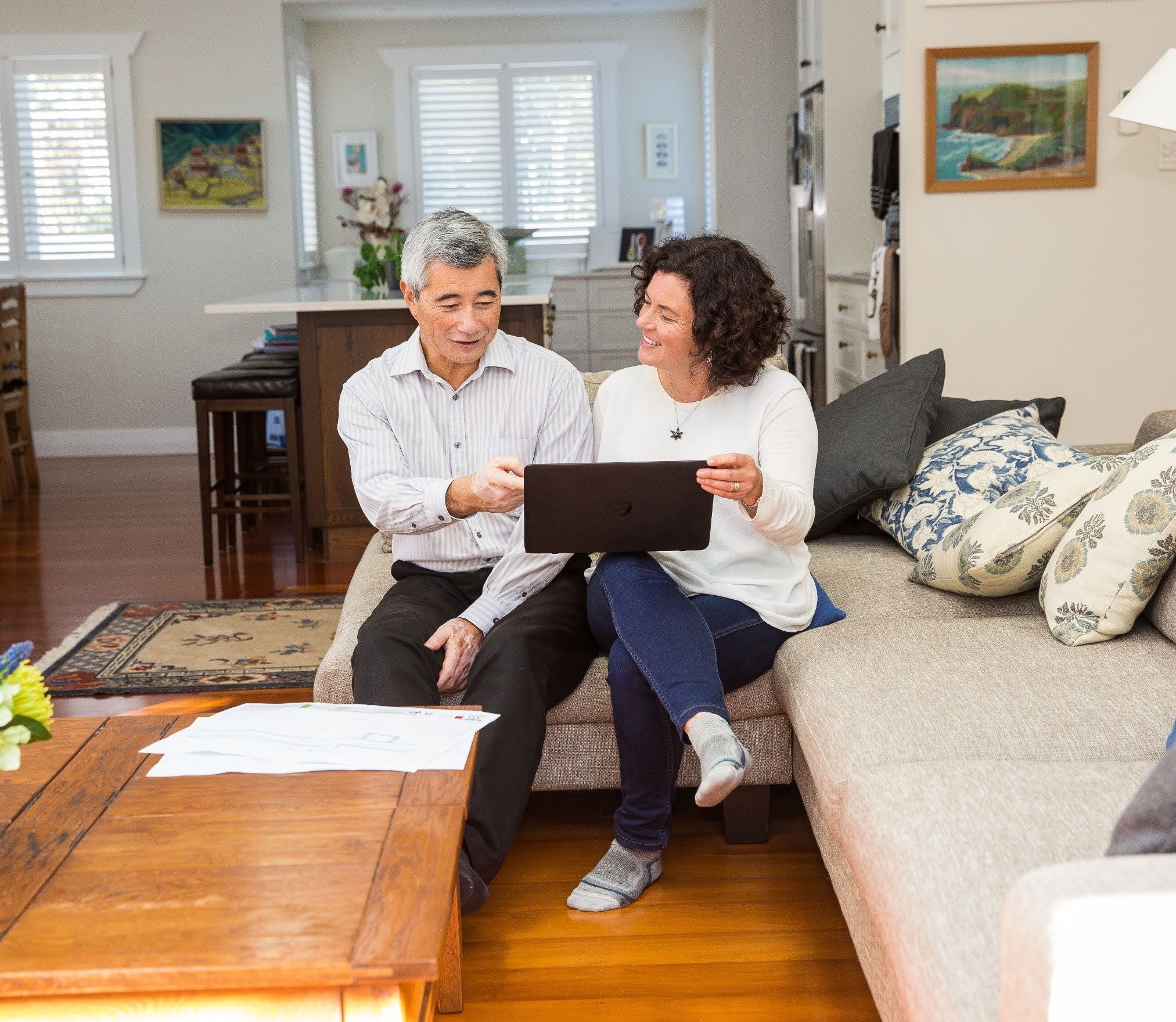 A couple happily chatting on a sofa at home looking at something on an ipad or laptop.