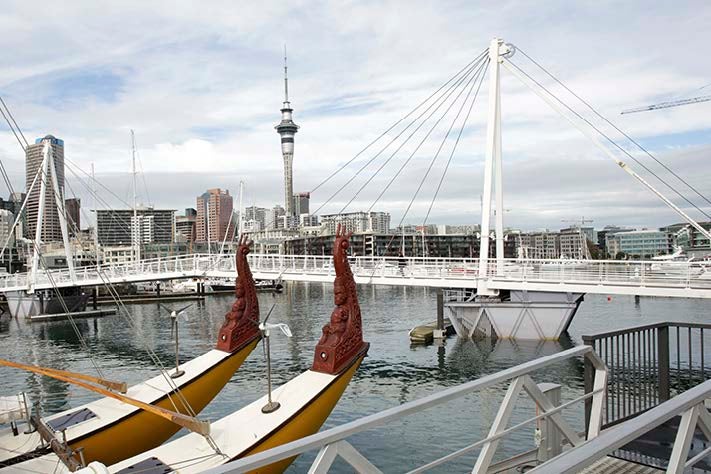 A double hulled, seagoing waka with beautiful carvings on the prow sits in the viaduct harbour, with the Auckland skyline in the background.