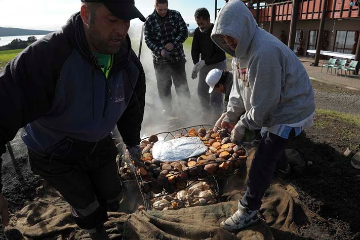 A group of folks are working on a hangi, taking out a basket of kumara out of the pit.