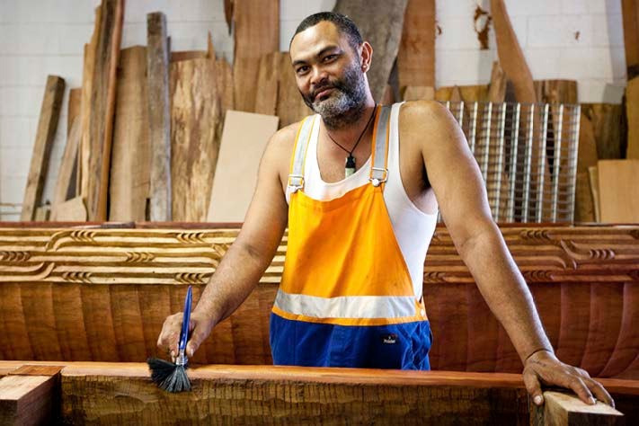 Local artist: Whare Thompson. He is a māori man with a beard, holding a paintbrush and in work clothes poses proudly infront of intricate, beautiful wooden carvings in his workshop.