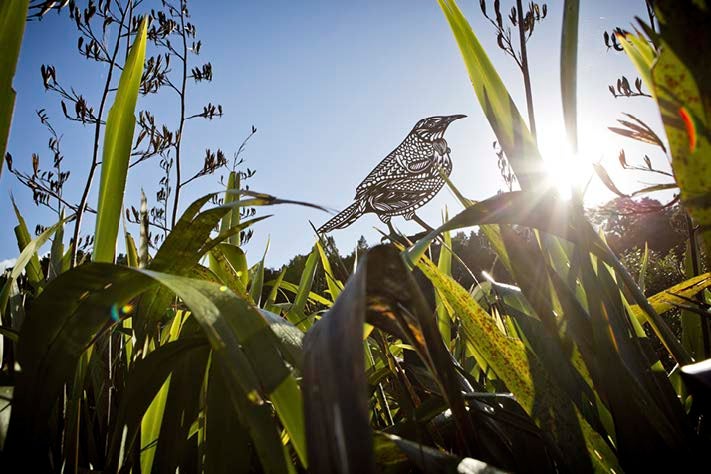 A laser-cutout of a tui bird overlooks a tussle of flax with flowers