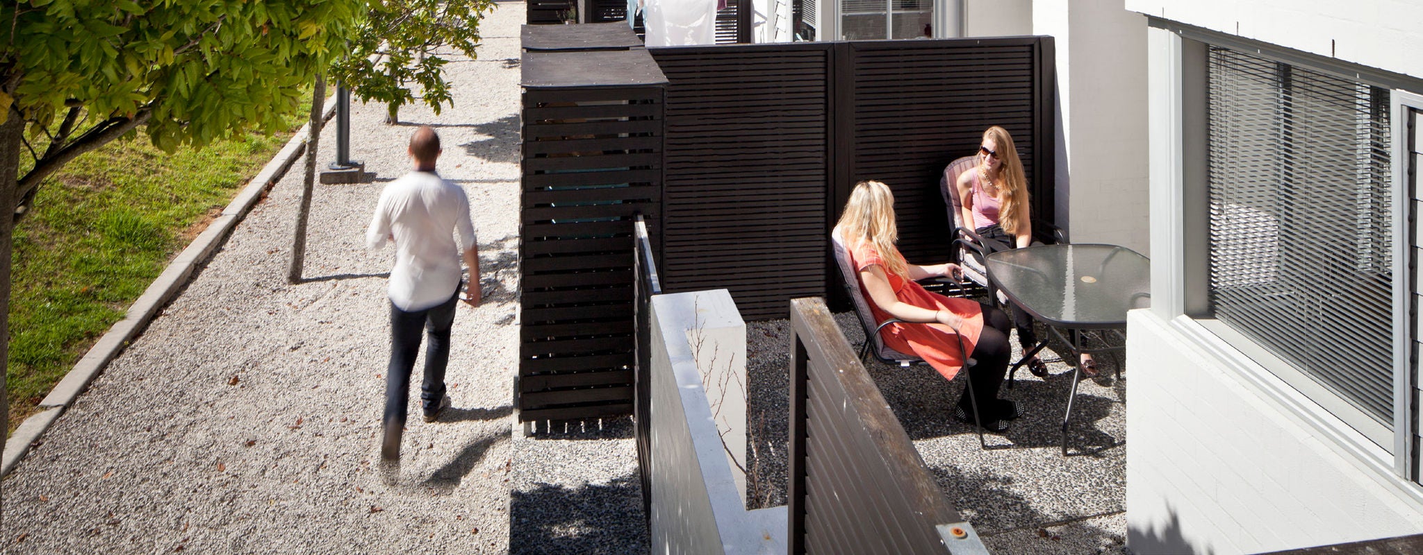 Someone is walking past on a sandy footpath as 2 women sit elevated on a balcony having a chat.