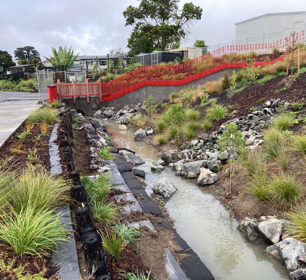 Te Ara Awataha greenway flows out of a culvert to a newly rehabilitated environment dotted with young native plants, rocks and embankment protections. These will make good homes for aquatic creatures native to the area. A stairway with a bright red balustrade flows over the top of the culvert, and in the background is Northcote primary school. It is a typical, partly cloudy Auckland day.